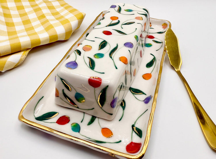Decorative butter dish with floral pattern on a white surface with a yellow checkered cloth and gold knife.